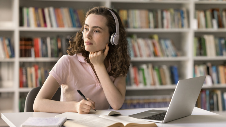 A woman listening to headphones in a library, next to a nearby laptop.