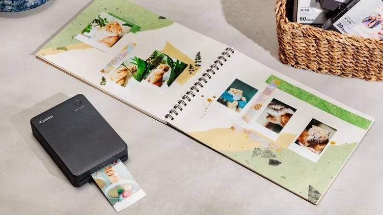 A scrapbook sitting next to the Canon Selphy QX20 instant printer, which is seen printing an image of a cat.