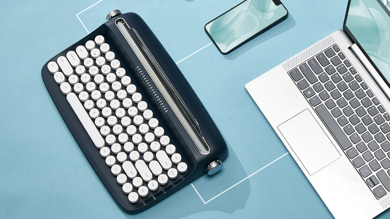 The Yunzii ACTTO B303 wireless typewriter keyboard on a blue surface, near a phone and a laptop.