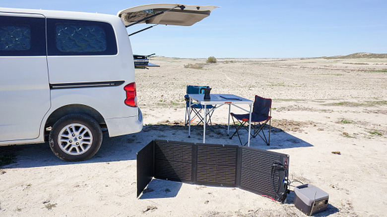 A folding solar panel being used to charge devices in a desert setting next to  a van with a small camp setup