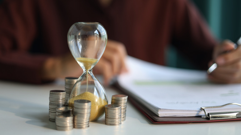 An hourglass surrounded by coins as a person works on a clipboard in the background