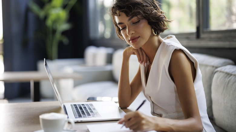 A woman at a table writing down notes next to her laptop