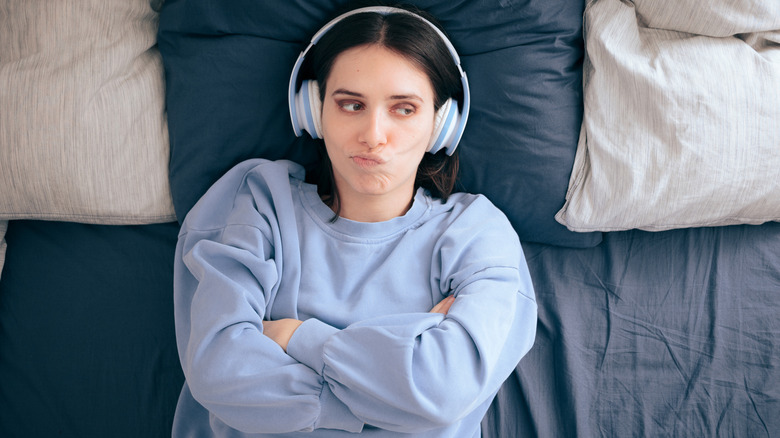 A woman wearing headphones lying on a bed with her arms crossed and an unhappy expression.