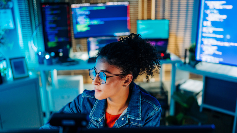 A woman sits in an office surrounded by computer monitors.