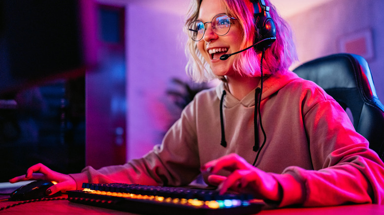 A woman looks at an offscreen gaming PC and smiles with her hands on the keyboard and mouse.