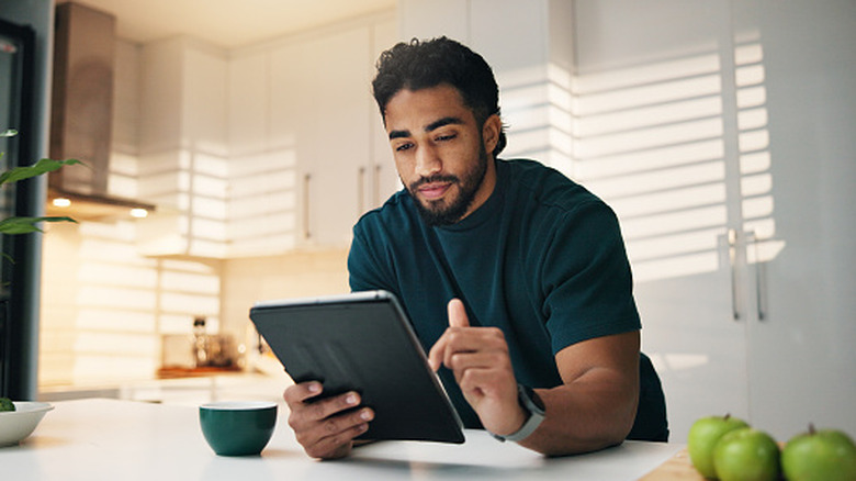 A man with a neutral expression using an iPad in a white kitchen.