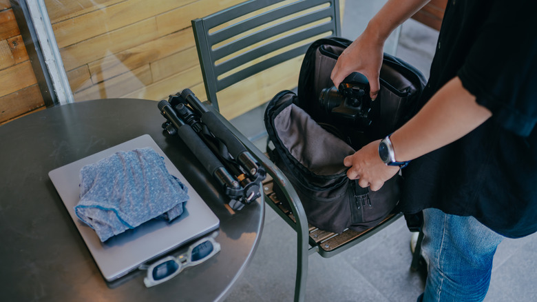 A person packing a camera into a padded photography backpack beside a table holding a folded tripod and a laptop. closed laptop, cloth, and sunglasses, preparing gear for a shoot.