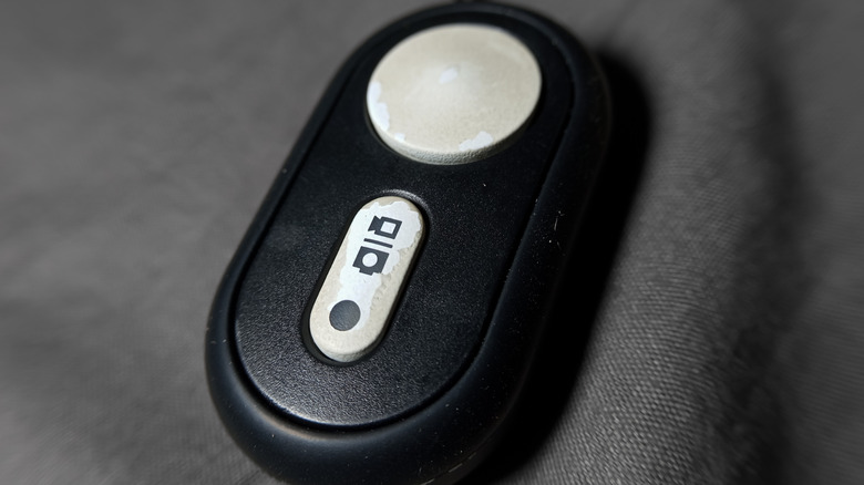 Close-up of a compact wireless camera remote shutter release with a large trigger button and camera icon, resting on a dark fabric surface.