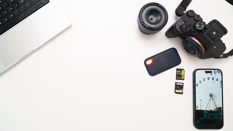 Photography gear on a white desk, including a mirrorless camera, lens, portable SSD, SD and microSD cards, smartphone, and laptop, arranged around a large blank workspace.