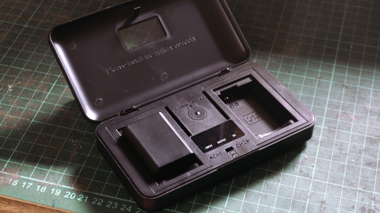An open dual-battery camera charger with one battery inserted and an empty charging bay resting on a cutting mat workbench.
