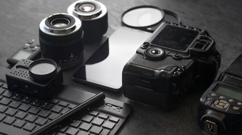 A collection of photography and content-creation equipment on a desk, including a mirrorless camera, interchangeable lenses, smartphone, keyboard, action camera, and wireless flash trigger.