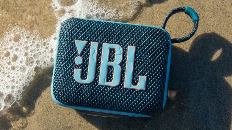 A blue JBL Go 4 Bluetooth Speaker with water droplets on it, placed on the sand and making light contact with water.
