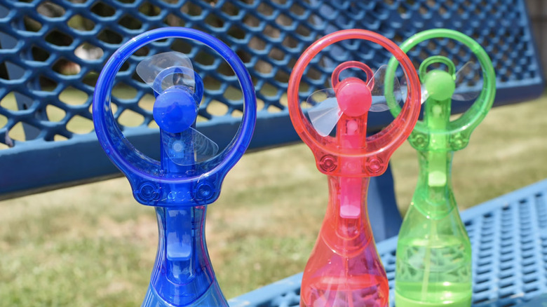 Three O2Cool Deluxe misting fans — one blue, one red, and one green — placed on a bench outdoors.