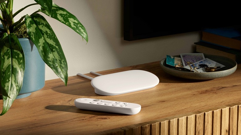 The Google TV Streamer streaming device on a table, below a TV and beside a plant.