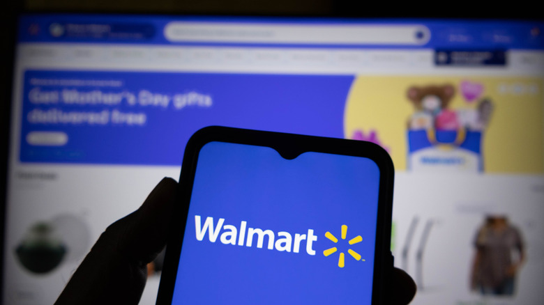 A person holding a smartphone showing the Walmart logo, with the Walmart website on a monitor in the background.