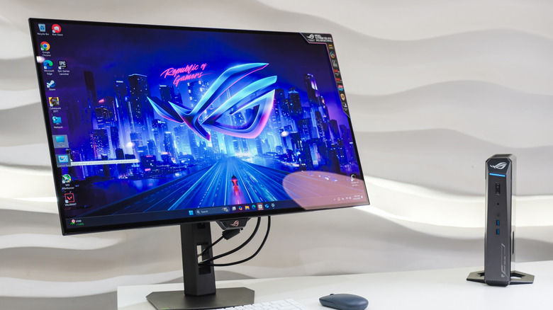 A black gaming monitor on a white desk displaying a cyberpunk-style Republic of Gamers desktop background