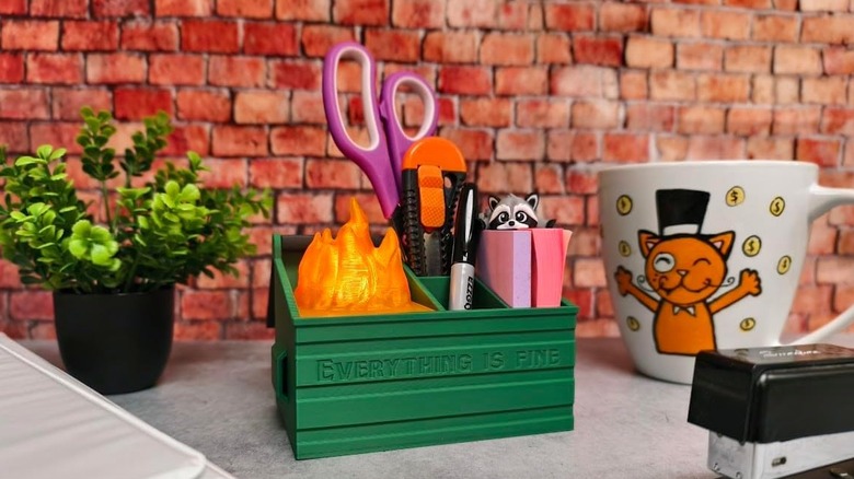 A green 3D-printed dumpster fire lamp and desktop tidy on a gray desk against a brick background