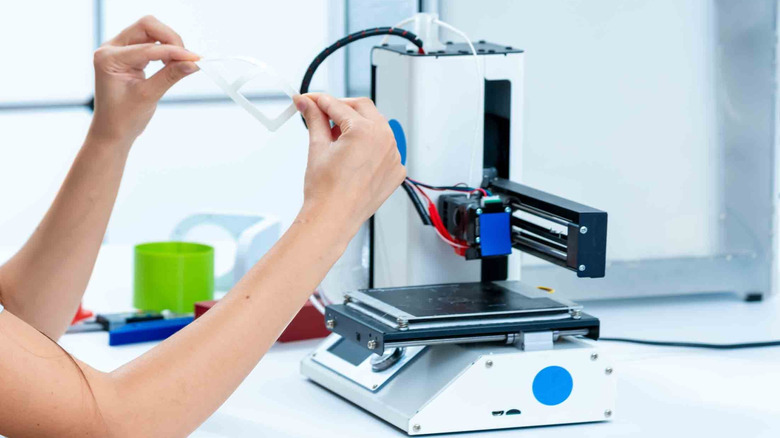 An open-bed 3D printer next to a person holding a 3D print.