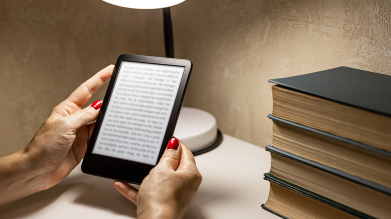 Hands holding an e-reader beneath lamplight next to a pile of books