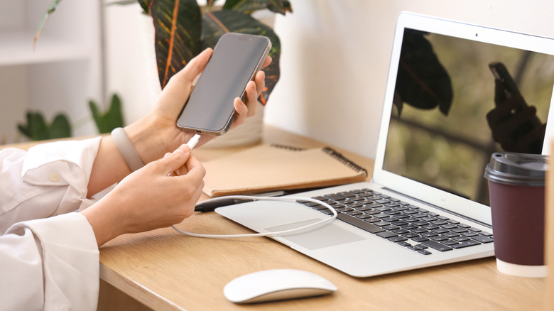 A person plugging in their phone to charge on a wooden desk, next to a Macbook, mouse, and a disposable coffee cup