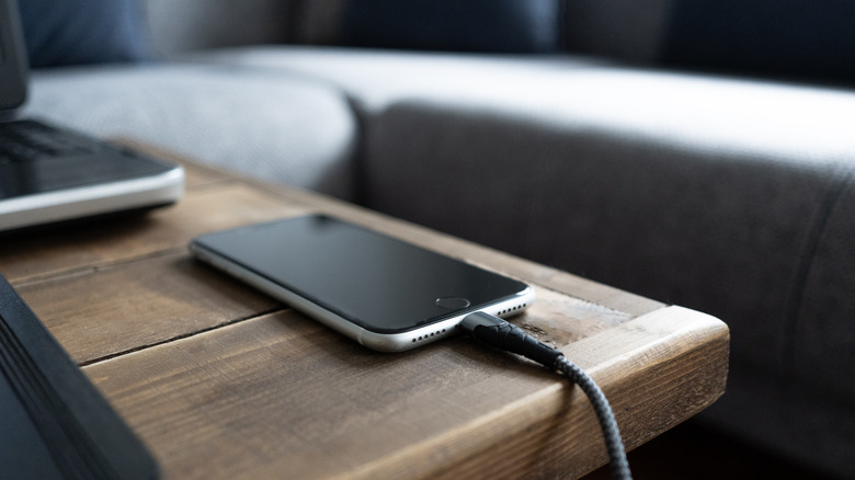 A phone plugged into a charger, placed on a wooden table that's near a couch