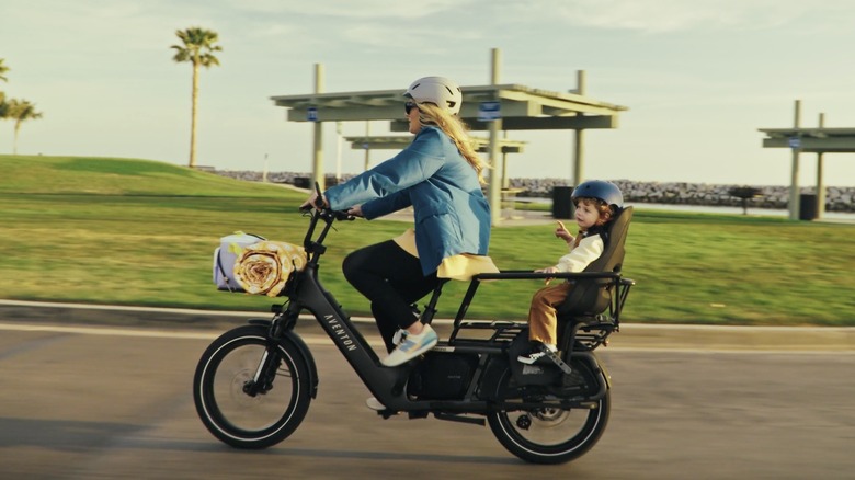 A woman riding on the Aventon Abound LR with child in the rear seat and picnic at the front. In the background are rolling grassy hills and palm trees.