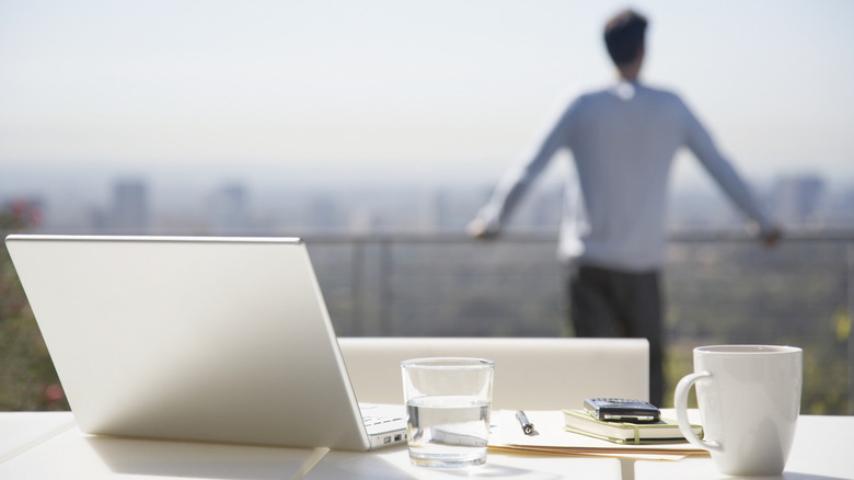 A person looking at a city skyline in the background, with their phone, notes, and laptop on a table in the foreground