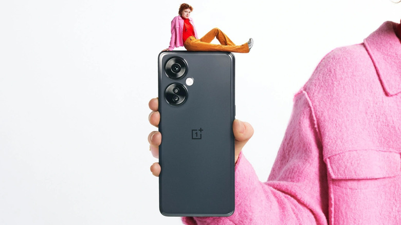 Gray OnePlus Nord N30 in a man's hand with a miniature human figure on top of it