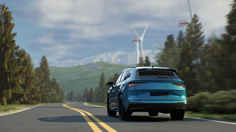 Modern EV driving along a green forest road with towering wind turbines.