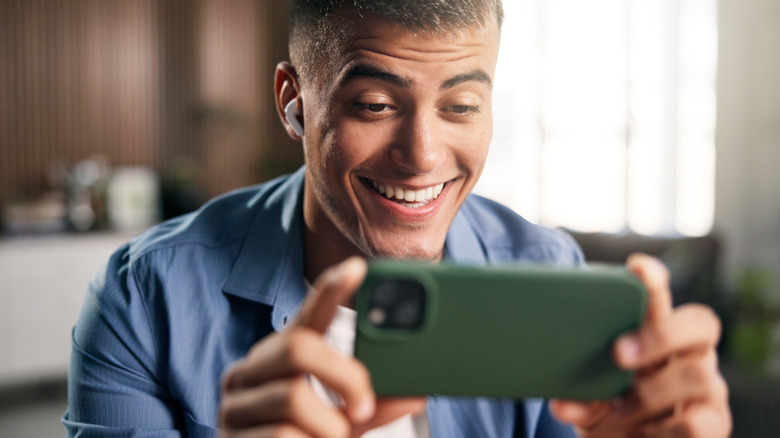 A man wearing earbuds and smiling while looking at his phone in landscape mode.