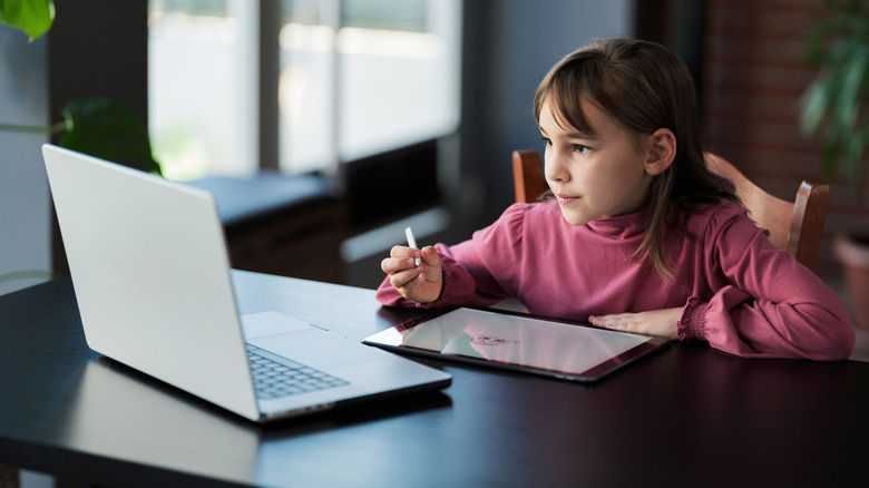 A young girl looks at a laptop screen while drawing on a tablet.