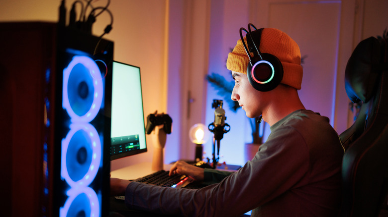 A PC gamer typing as his computer wearing headphones and an orange beanie.