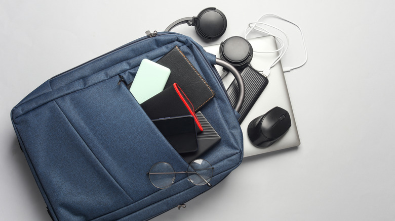 A silver laptop and various accessories, including a mouse, power bank, and headphones, spilling out of a navy backpack.