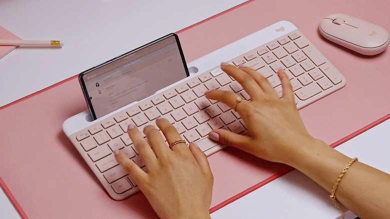 A person typing on a rose-colored Logitech K585 keyboard with a smartphone placed in its phone holder slot.
