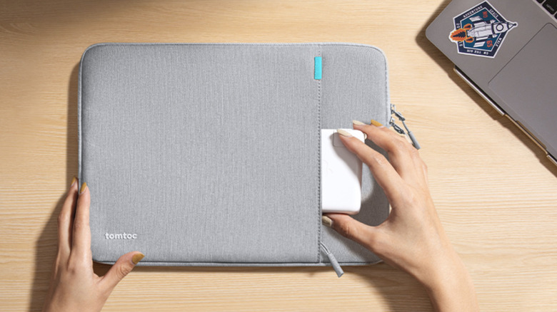 A person placing a MacBook charger base into the slot of a gray Tomtoc 360° Protective Laptop Sleeve.