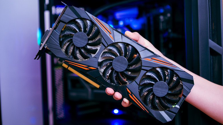 A hand holds a large triple-fan graphics card in front of an open PC case.