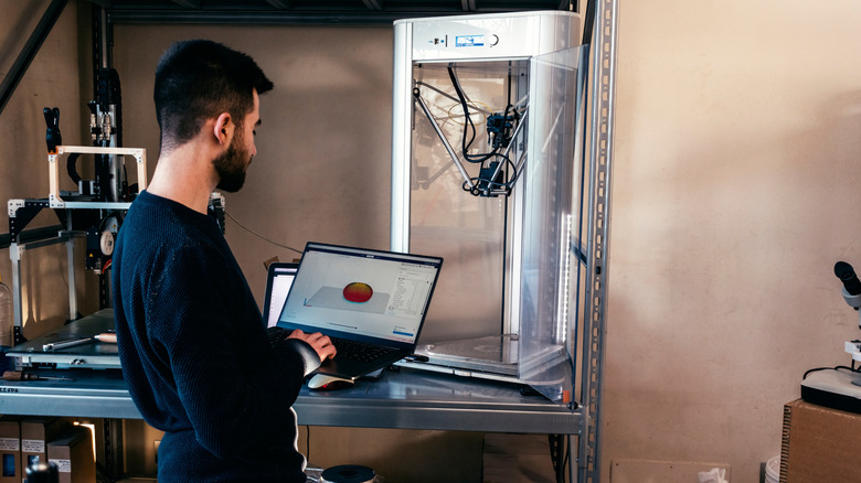 A person holding a laptop stands next to a working 3D printer.