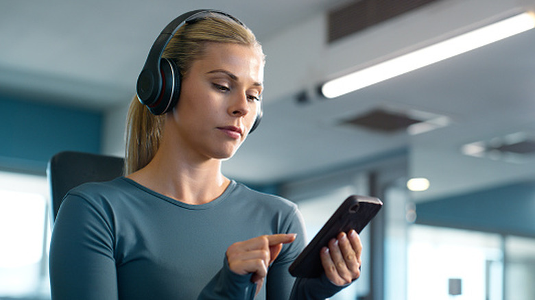 A woman in a gym wearing headphones operates her smartphone.