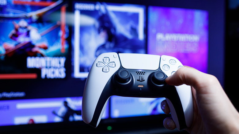 A hand holding a black and white PS5 controller in front of a blurred PS5 store page