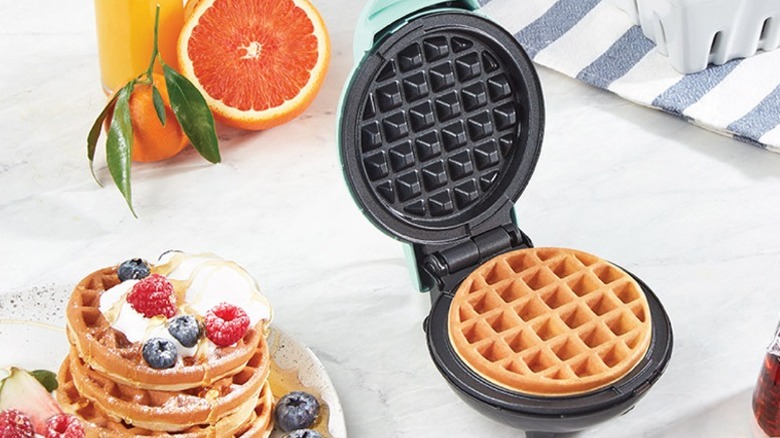 Waffle placed inside a waffle maker alongside oranges and a plate of served waffles.