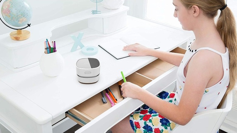 Girl sitting on a study table with the vacuum cleaner placed on the table and drawers open.