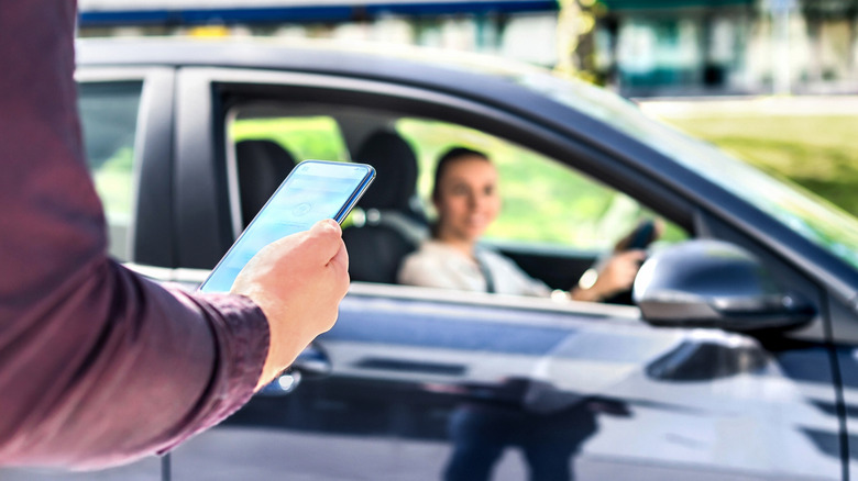 A person checking their phone in front of a car with a driver smiling at them in the background