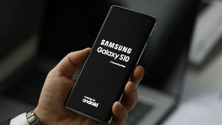 Male hand holding Samsung Galaxy S10 Plus with a laptop in background