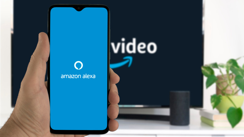 Hand holding a mobile with the Amazon Alexa app icon with a TV displaying Prime Video logo
