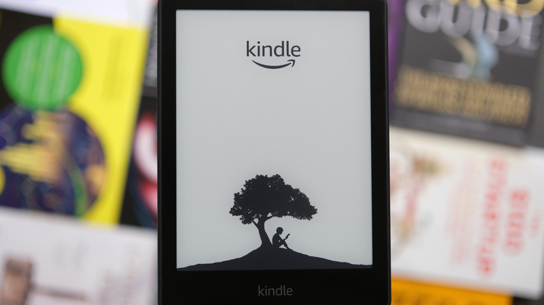 A close-up of a Kindle reader with blurred books in the background.