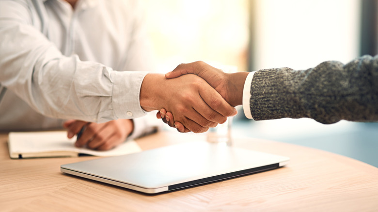 Two individuals shaking hands over a closed laptop sitting on a table
