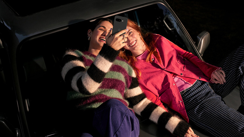 Two women sitting on the hood of a car with a Google Pixel 9a.
