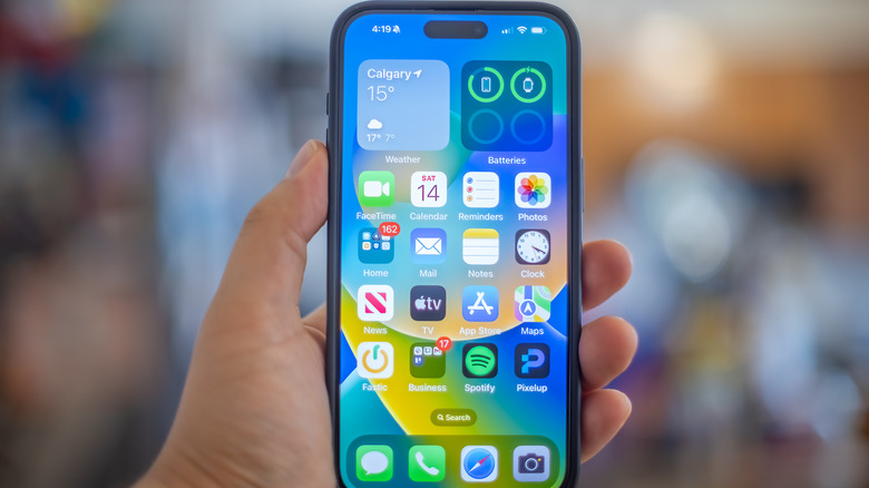 A hand holding an iPhone with the home screen displayed.