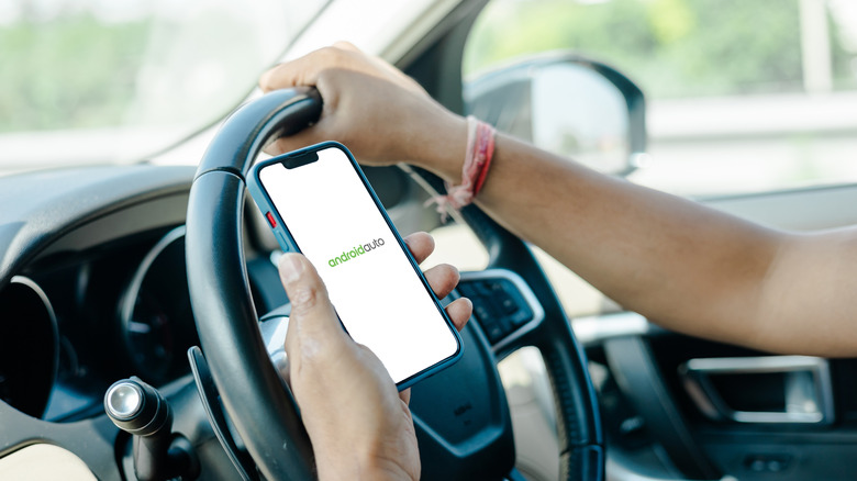 Person holding phone in one hand and steering wheel in other, with Android Auto logo on screen
