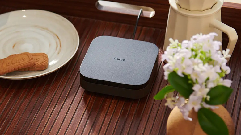 Aqara Hub M3 smart home hub on a wooden table beside a plate and flowers.